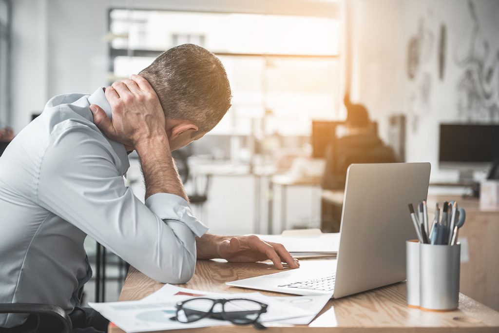 Man holding sore neck while using notebook computer with headache. Needing headache treatment.