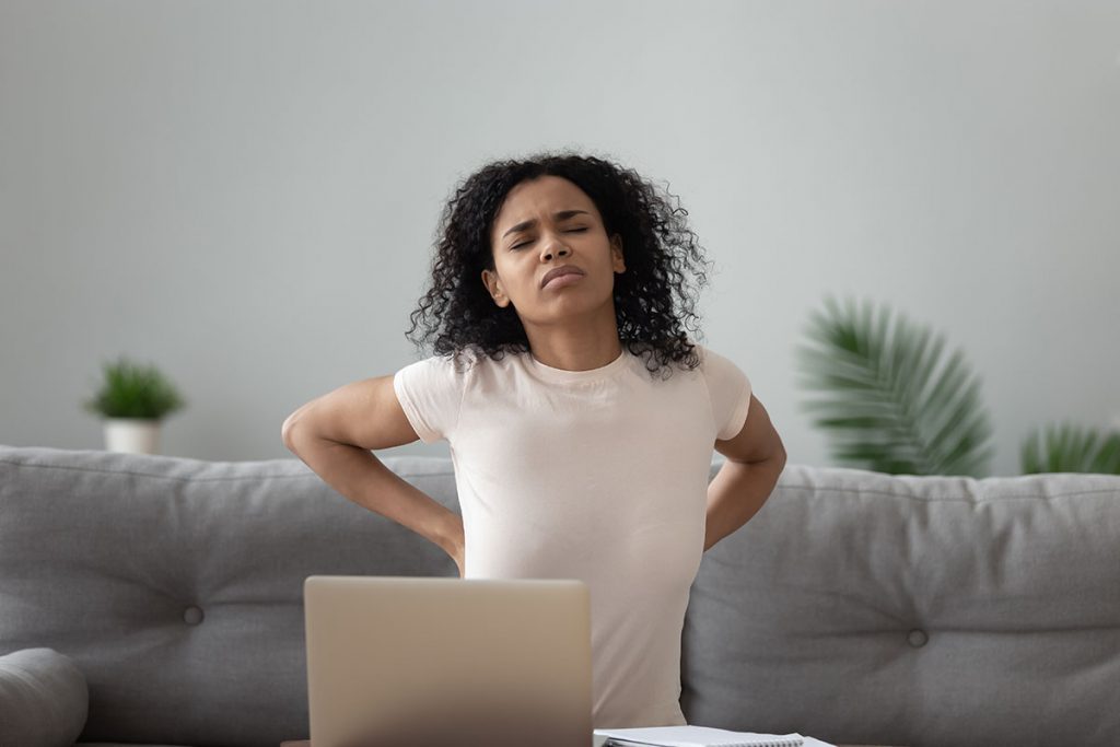 Woman sitting on sofa touches lower back suffers from severe pain caused by overuse of computer lot of time working on pc, poor posture and improper spinal support concept